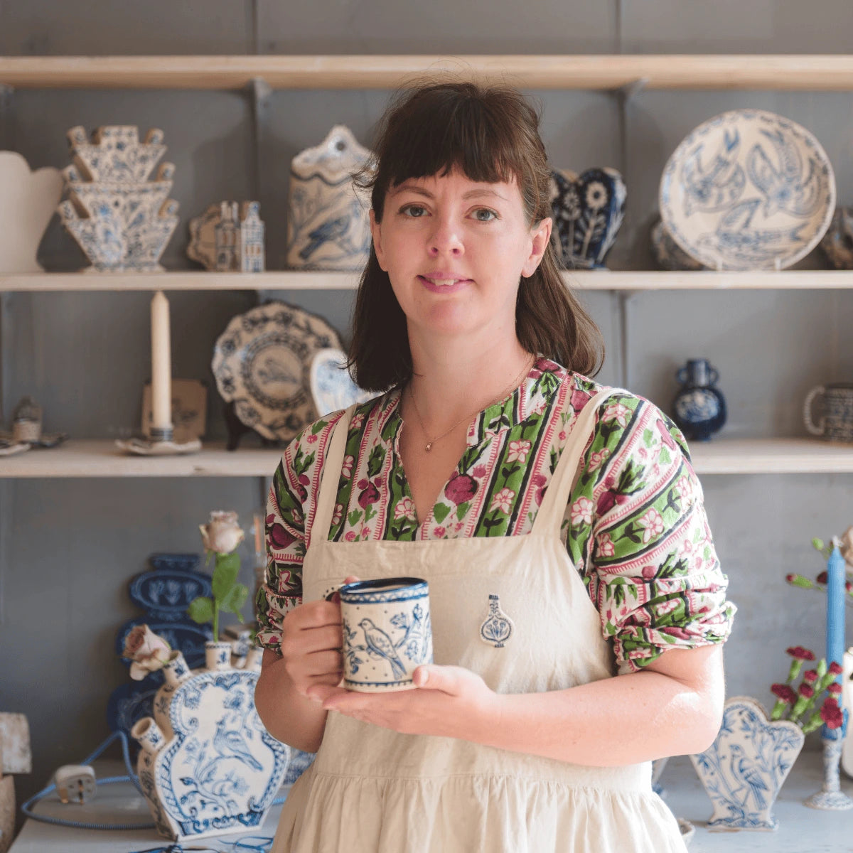 Woman holding a ceramic mug in a pottery studio with shelves of ceramic items.