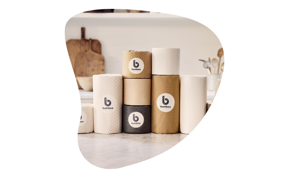The image shows a kitchen setting with a clean, minimalist design. In the foreground, there is a variety of paper products, including toilet paper rolls, paper towels, and tissues, all from the brand "bumboo." The products are arranged neatly on a marble countertop. Each product is wrapped in eco-friendly packaging featuring natural colors like beige, brown, and black, with the brand logo prominently displayed. In the background, the kitchen has a white theme with wooden elements, including cutting boards