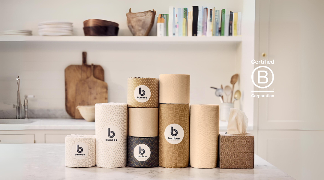 A collection of bamboo paper products from the brand "bumboo." The items are arranged in a neat row on a kitchen countertop. The collection includes various types of rolls and a box of tissues, all in natural, unbleached colours. Each product features the "bumboo" logo, which is a stylised letter "b" inside a circle, prominently displayed on the front. The background includes a wooden cutting board and some kitchen utensils, giving a warm, homey feel to the scene.