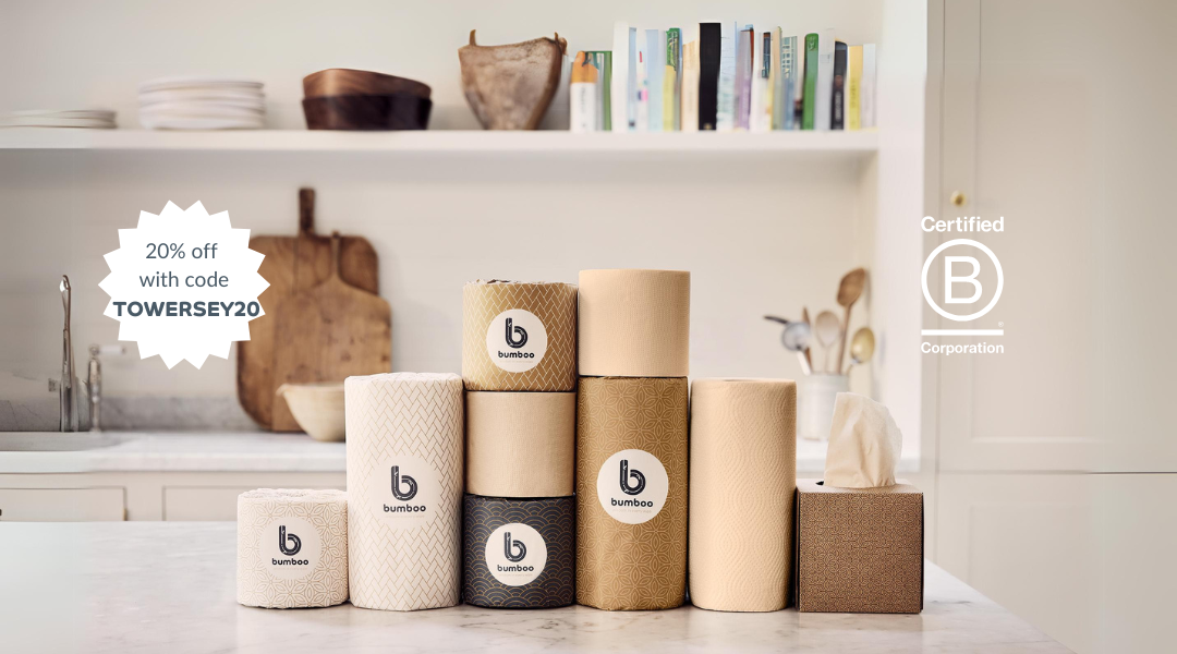 itchen setting with a clean, minimalist design. In the foreground, there is a variety of paper products, including toilet paper rolls, paper towels, and tissues, all from the brand "bumboo." The products are arranged neatly on a marble countertop. Each product is wrapped in eco-friendly packaging featuring natural colors like beige, brown, and black, with the brand logo prominently displayed.