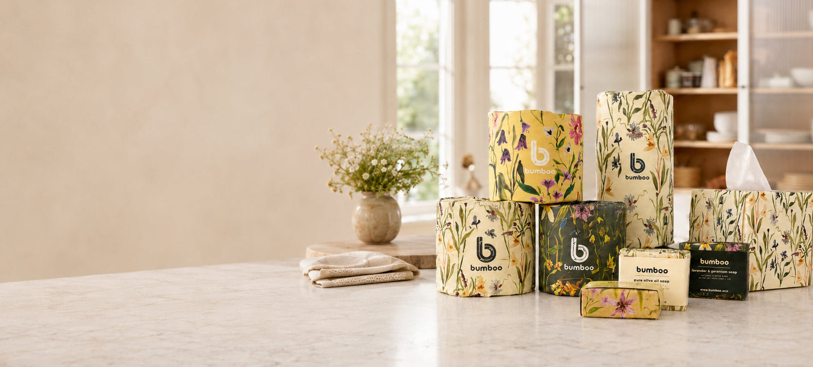 A group of bamboo tissue boxes and tissue packs with floral-patterned yellow and green packaging stands on a light marble countertop in a bright, modern kitchen setting.