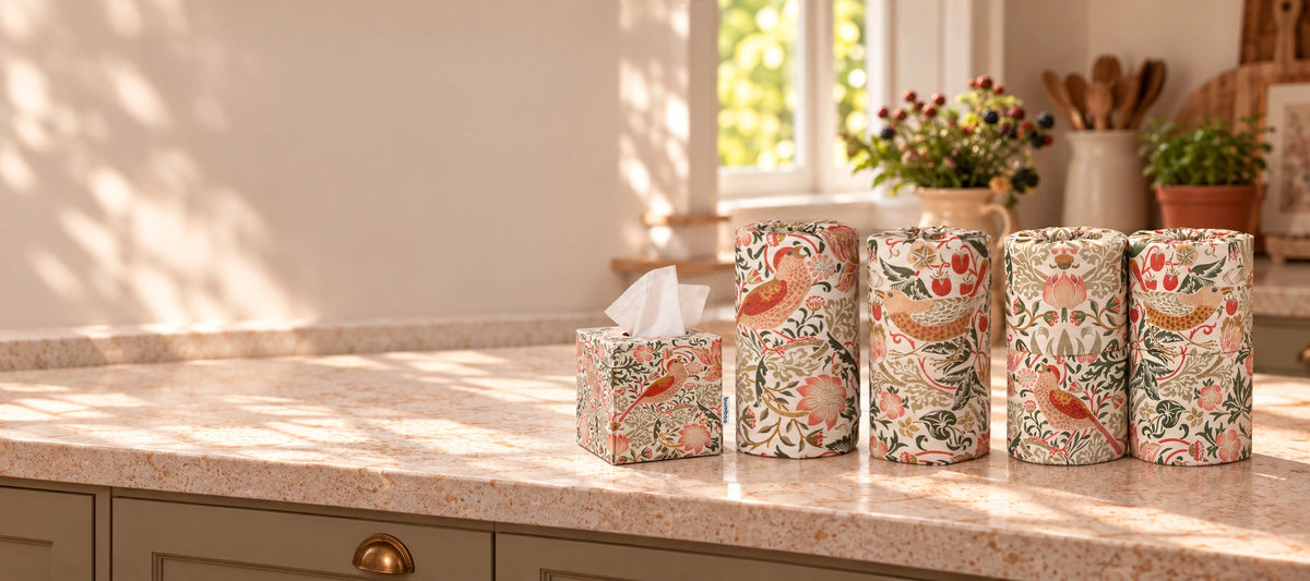 Four decorative tissue boxes and matching toilet rolls and kitchen rolls with a William Morris-style floral and bird pattern in muted pink, green, and beige tones, arranged on a sunlit kitchen countertop.