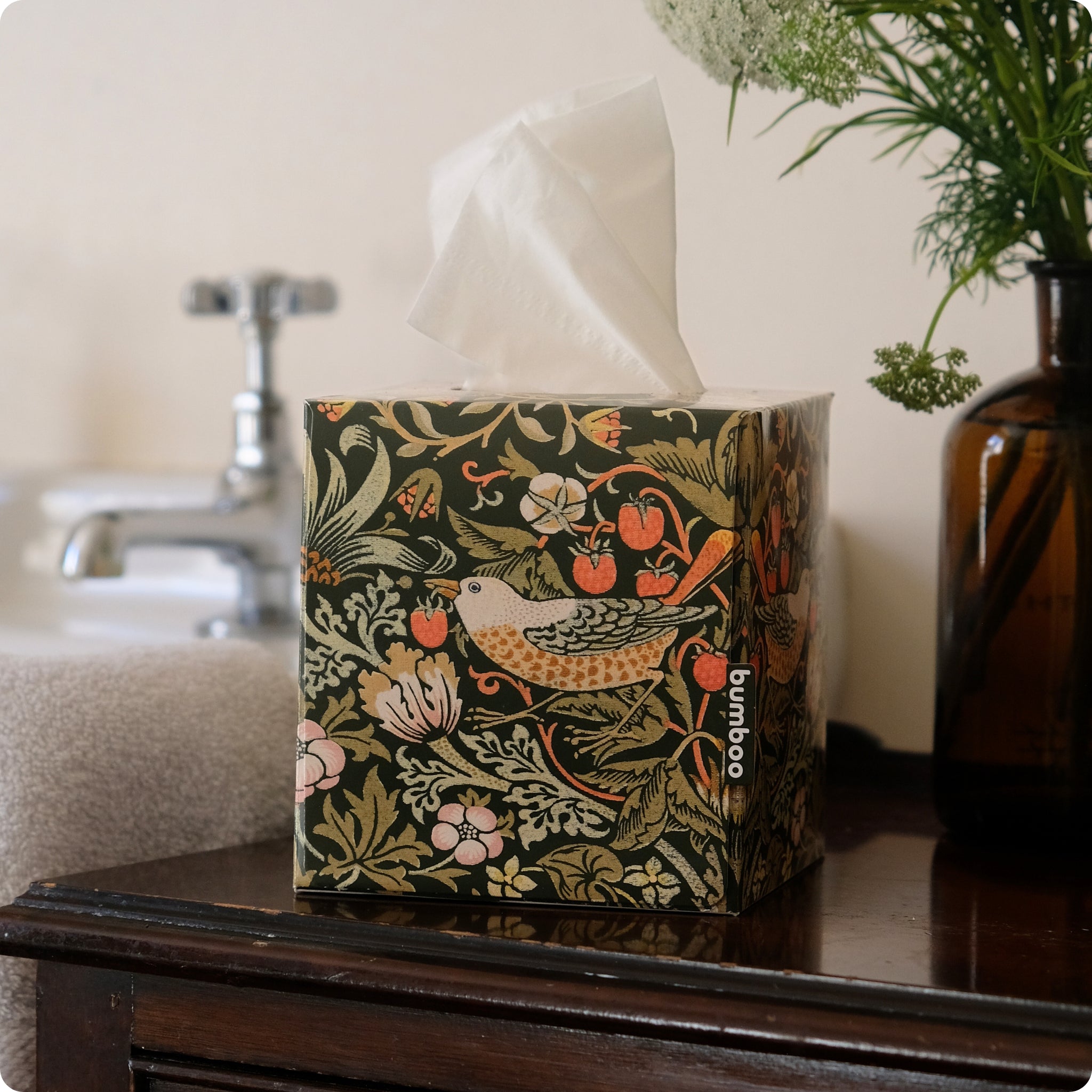 A tissue box cover with a dark green William Morris-style floral and bird pattern sits on a dark wooden table in a bathroom setting, with a tissue pulled from the top. The brand name 'bumboo' is printed vertically on the side.