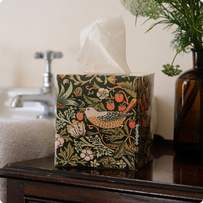 A tissue box cover with a dark green William Morris-style floral and bird pattern sits on a dark wooden table in a bathroom setting, with a tissue pulled from the top. The brand name &