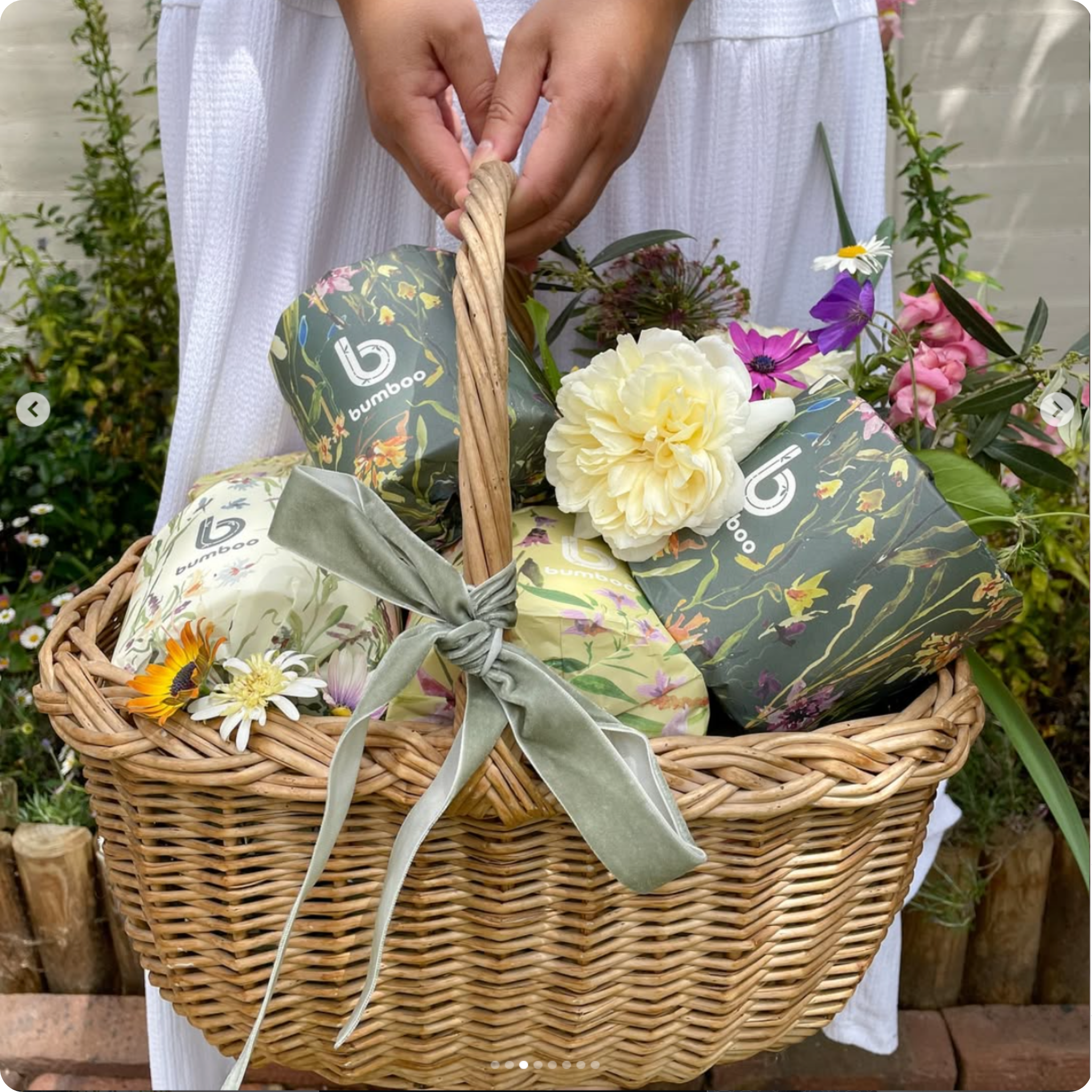 Wicker basket with floral arrangements and Bumbaa products, held by a person in a white dress.
